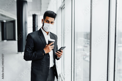 A young Indian businessman in a protective mask by the window, he holds his phone in his hands and looks at the camera