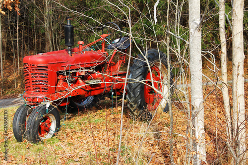 A small, old fashioned, red farm tractor parked along side a country road, among the trees, autumn. The tractor has been strung with Christmas lights.