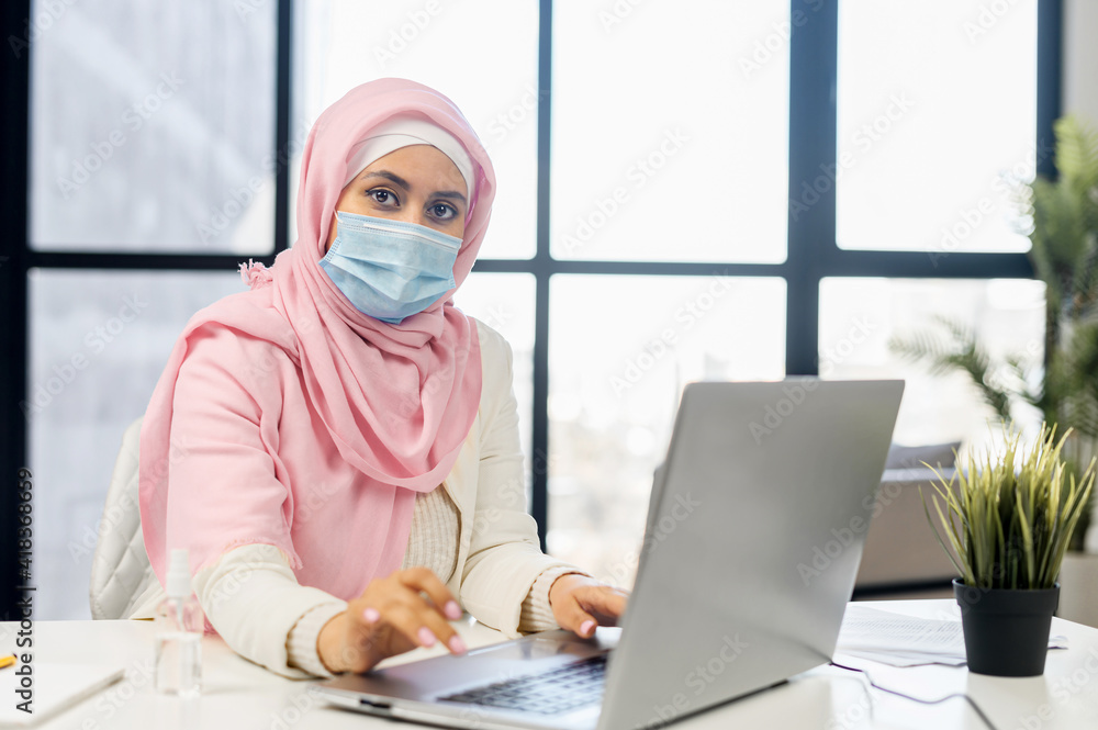 Serious muslim woman wearing hijab and protective face mask using laptop in modern office space. Islamic businesswoman in surgical mask looks at the camera sitting at the desk Serious muslim woman wearing hijab and protective face mask using laptop in modern office space. Islamic businesswoman in surgical mask looks at the camera sitting at the desk