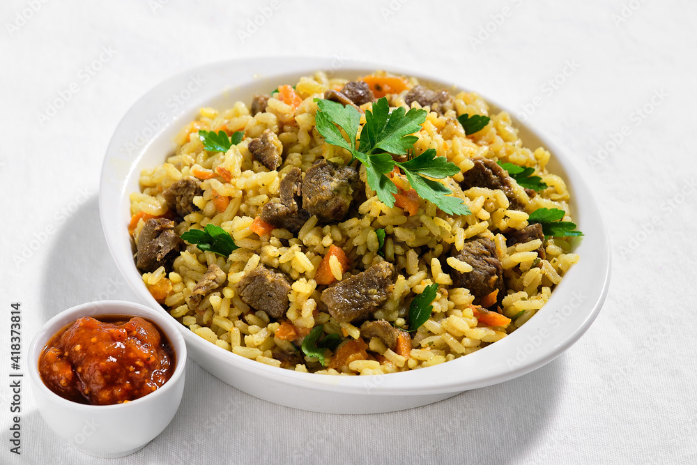 Plov, pilaf or pilau, rice dish from Central Asia made with golden rice, carrot and browned lamb meet. The portion is served in ceramic bowl on off white textile tablecloth. Ajika sauce on the side.