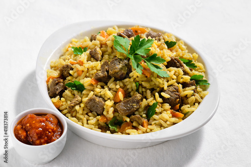 Plov, pilaf or pilau, rice dish from Central Asia made with golden rice, carrot and browned lamb meet. The portion is served in ceramic bowl on off white textile tablecloth. Ajika sauce on the side.