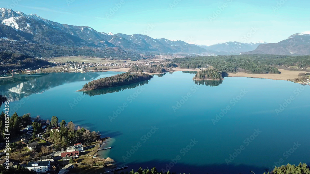 A drone shot of a Faaker lake in Austrian Alps. The lake is surrounded by high mountains. There is a small island in the middle. Green forest growing at the shore. Clear and sunny day. Calmness
