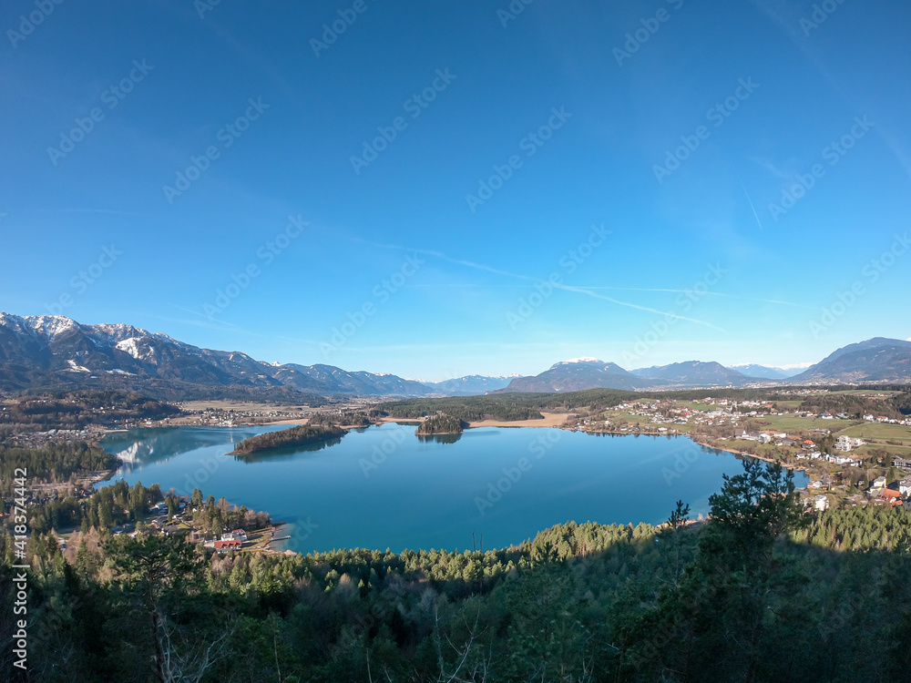 A drone shot of a Faaker lake in Austrian Alps. The lake is surrounded by high mountains. There is a small island in the middle. Green forest growing at the shore. Clear and sunny day. Calmness