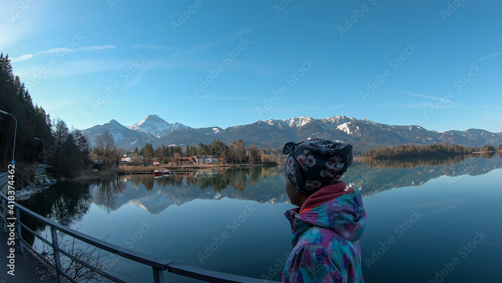 A woman in winter jacket walking along the shore of Faaker lake in Austrian Alps. The lake is surrounded by high mountains. Calm surface of the lake reflects the surrounding. Clear blue sky. Serenity