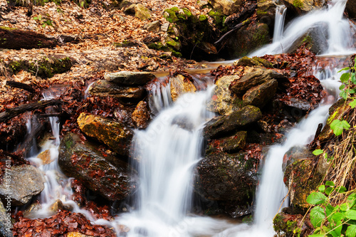A landscape with a small waterfall surrounded by dry leaves and mosses on the rocks, whose flowing water was photographed in the foreground, creating a blurred effect