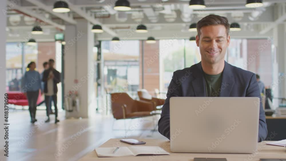 Dolly shot of casually dressed businessman working on laptop taking coffee break at desk in modern open plan start up office with colleagues in background - shot in slow motion Dolly shot of casually dressed businessman working on laptop taking coffee break at desk in modern open plan start up office with colleagues in background - shot in slow motion