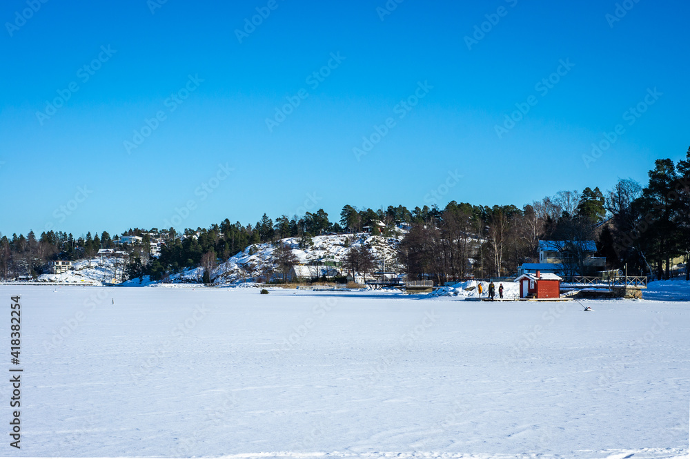 Scandinavian winter amazing  landscape. Frozen Baltic sea bay in Northern Europe. People stand on the pier near small red wooden house cabin or bathhouse. Rocky shores growned with evergreens. 