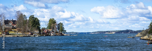 Panoramic view of the Baltic Sea Bay on sunny spring day. Rocky shores of Scandinavia covered with evergreen forests. Traditional Swedish red wooden houses on the coast. Beautiful white clouds on sky.