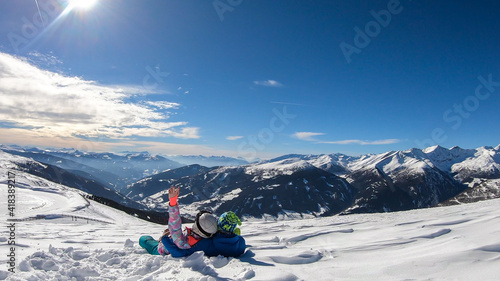 A couple sitting on the snow on top of Katschberg in Austria. They are happy and joyful. Panoramic view on the surrounding mountains. Winter wonderland. Bright and sunny winter day. Love and happiness