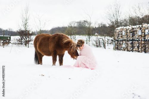 Eine wunderschöne Frau im Rosa Tüll Kleid, sieht aus wie Aschenputtel, steht  im Schnee und füttert mit ihren Pony