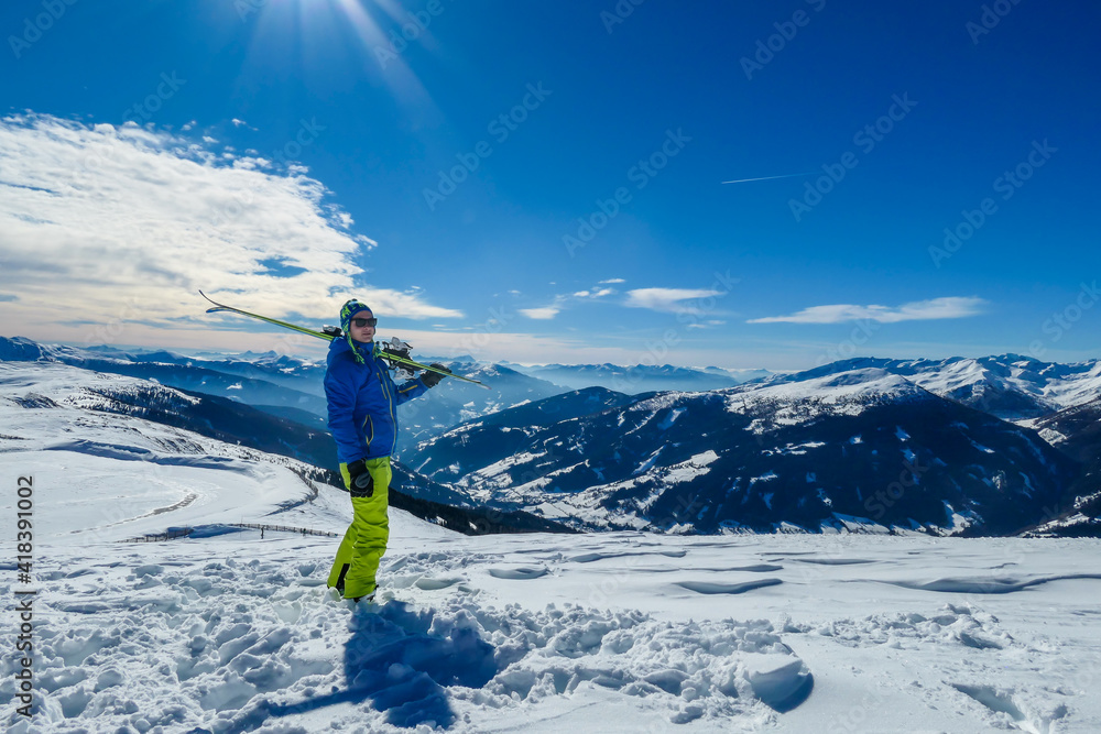 A man posing on powder snow with his skies on top of Katschberg in Austria. Panoramic view on the surrounding mountains. Winter wonderland. Sunny winter day. He is full of energy and happy. Adventure