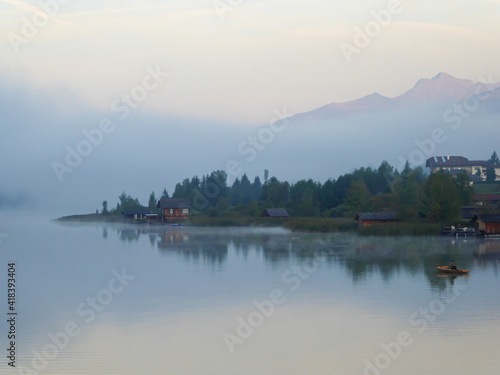 A foggy morning at the Weissensee lake in Austria. The lake is surrounded by high Alps. The calm surface of the lake reflects the forest. Sunbeams reaching the high peaks. Serenity and peacefulness