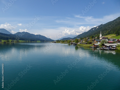 An idyllic, panoramic view on the Weissensee lake in Austria. The lake is surrounded by high Alps. There is a small village at the lake's shore, with tall church tower. Few clouds. Calmness and peace