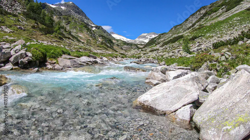 A rushing torrent in the Austrian Alps. The meadow around it is overgrown with lush green grass. In the back there is a glacier. Sunny and bright day. Power of the nature. Remedy and serenity