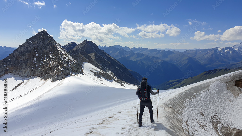 A man with hiking stick hiking through a glacier in Heiligenblut region in Austria, on the way to Hohe Sonnblick. There are many high chains of Alps behind him. Sunny day. Adventure and discovery.