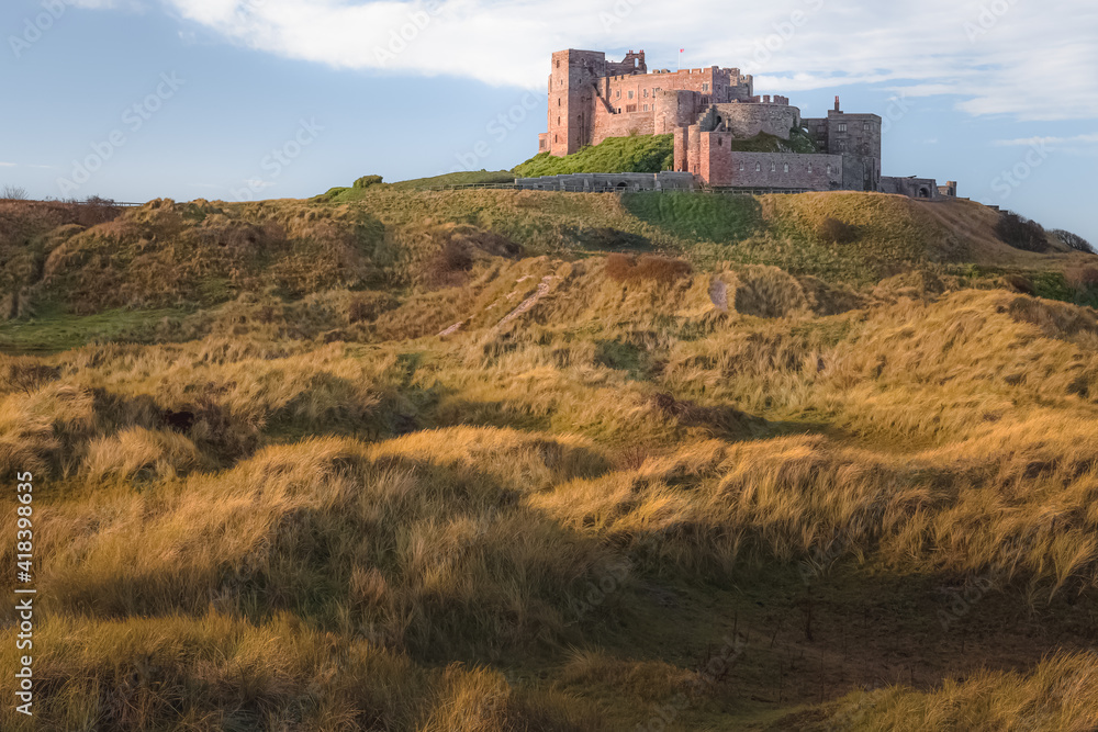 View of the historic Bamburgh Castle from the long grassy dunes in golden light on the northeast coast of England, Northumberland.