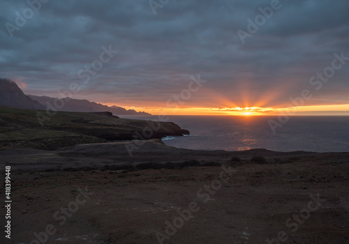 Beautiful sunset on the rocky atlantic coast in north west part of Gran Canaria island. Red orange sun with rays going down to the ocean. Dark blue clouds and cliffs in background.