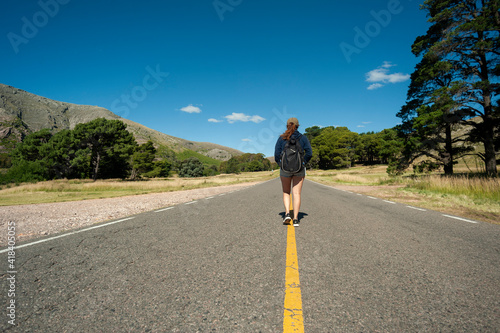 Young Latin woman hitchhiking on the road with a backpack waiting for someone to give her a ride in the mountains and forests on a sunny day.