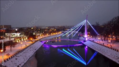 Bridge (Mist Zakokhanykh) across river in blue purple light illumination with beautiful reflection, Skver Strilka in Kharkiv city center. Winter aerial evening colorful footage photo