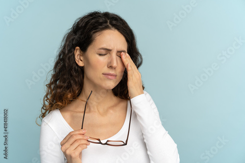 Sleepy young caucasian woman holding glasses rubbing eyes, feels tired after working on computer, isolated on blue background. Exhausted office employee suffering from ocular diseases, eye strain. 