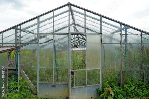 Old large greenhouse for agricultural cultivation, now out of use with some broken glass panes and overgrown with weed and tall grass. 