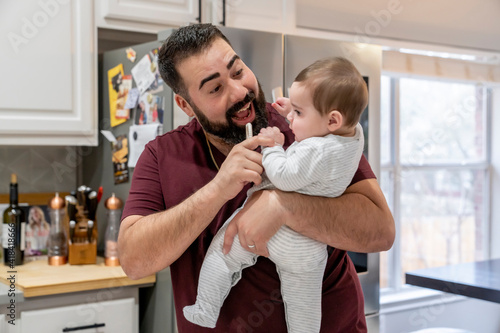 Man with beard holding and tickling baby in kitchen photo