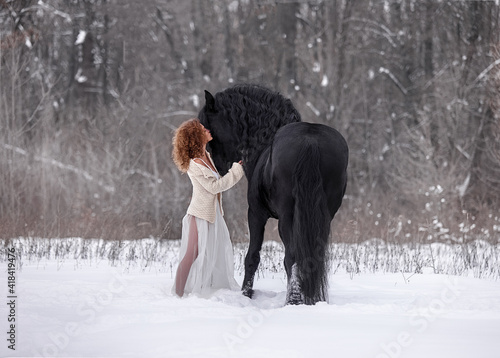 A beautiful curly-haired girl stands next to a Friesian stallion with a long mane