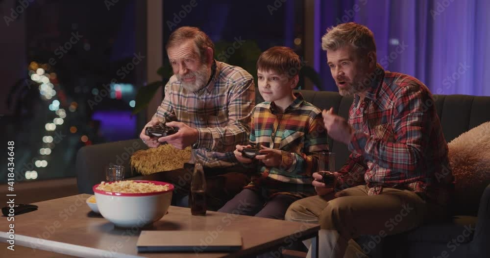 Family of three playing computer games on console together. Multi-generation trio father son and grandpa having fun at home weekend night. Family of three playing computer games on console together. Multi-generation trio father son and grandpa having fun at home weekend night.