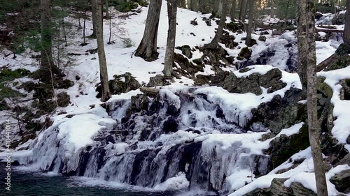 A winter waterfall cascades down layers of rock and ice into the river below.