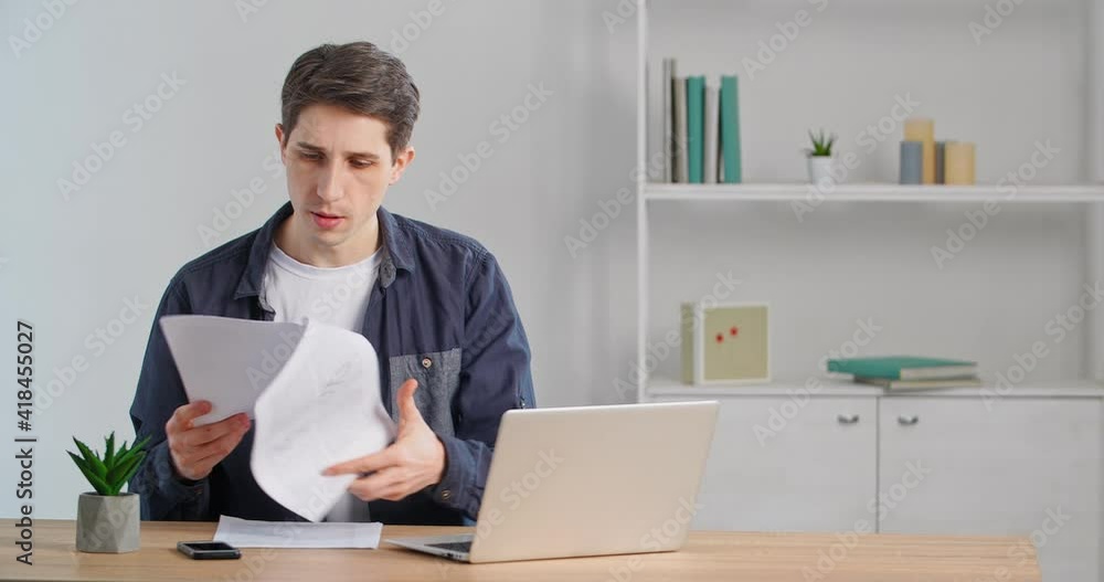 Caucasian male freelancer sits at table in his home office reads documents compares numbers in papers and enters data into bank payment service in laptop calculates budget using financial app online Caucasian male freelancer sits at table in his home office reads documents compares numbers in papers and enters data into bank payment service in laptop calculates budget using financial app online
