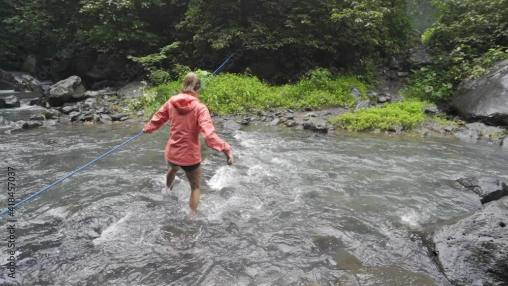 Slow motion lockdown with a pan right of a woman hiker carefully crossing a rushing river with the help of a guide rope, all surrounded by rocks and tropical plants - Bali, Indonesia Slow motion lockdown with a pan right of a woman hiker carefully crossing a rushing river with the help of a guide rope, all surrounded by rocks and tropical plants - Bali, Indonesia