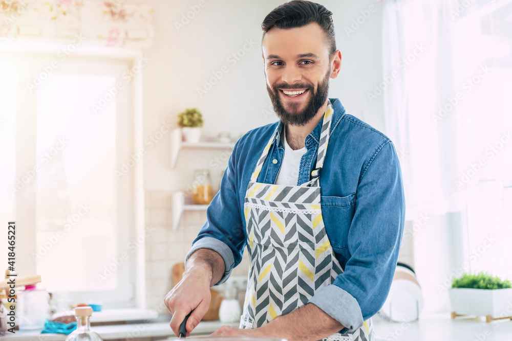 Handsome smiling young bearded man cooks fresh healthy vegan salad for his family and having fun in the big light kitchen