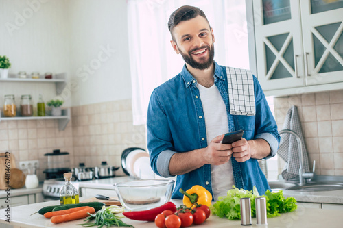 Happy handsome bearded man is using his smart phone while he preparing vegan healthy breakfast for a lovely family