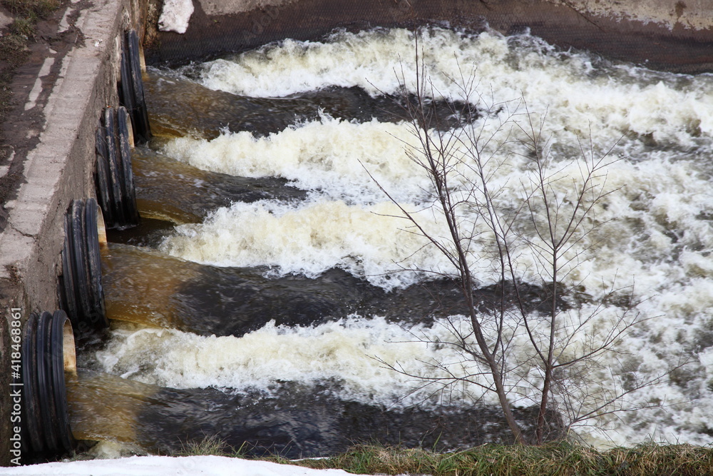 Closeup powerful fast bubbling water stream from a storm drain spillway pipes on dry bare tree