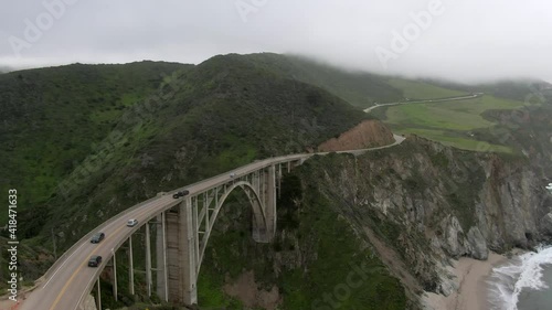 Aerial shot of vehicles on Bixby Creek Bridge by sea, drone flying forward towards green landscape during foggy weather - Big Sur, California