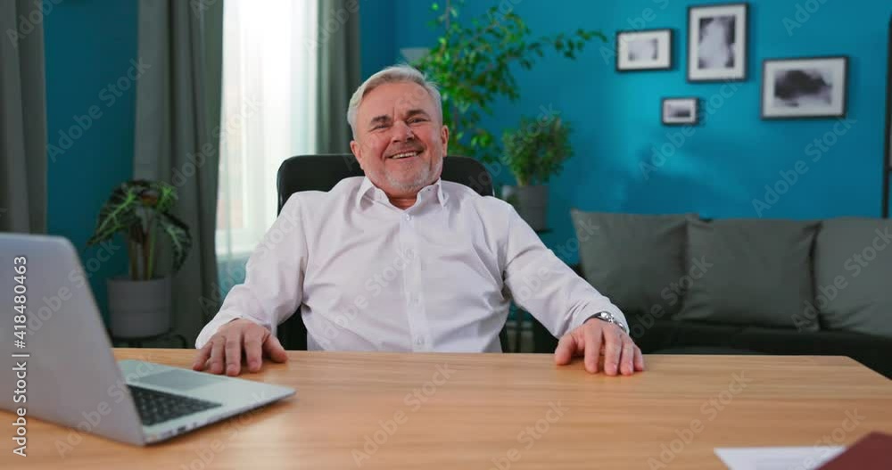 Professional mature director sitting at his desk in his home office studio working on his laptop is happy and celebrating business success by dancing, singing and happily raising his hands up. Professional mature director sitting at his desk in his home office studio working on his laptop is happy and celebrating business success by dancing, singing and happily raising his hands up.