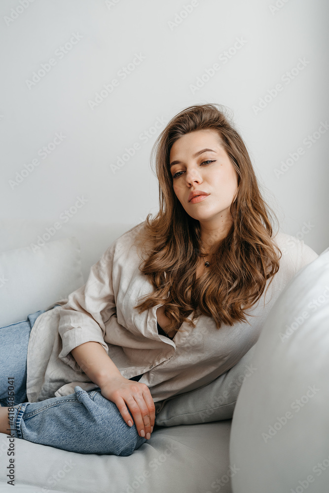 A young blonde woman in casual comfortable clothes sits on a white sofa at home. scandinavian interior, minimalism A young blonde woman in casual comfortable clothes sits on a white sofa at home. scandinavian interior, minimalism