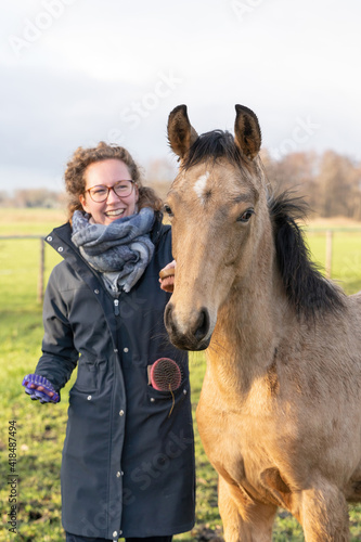 Smiling young woman in a raincoat with her yellow 1 year old stallion in the pasture. Curious horse's head while being brushed. Selective focus