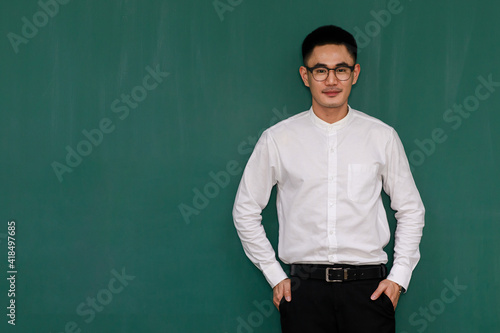 Portrait of young and handsome Asian man wearing eyeglasses and casual business clothes, white shirt and black trousers, standing pose with self-confidence with green background and copy space