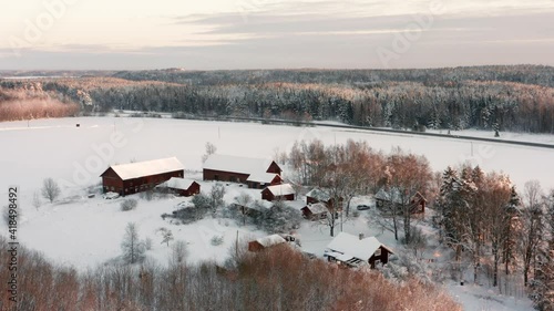 Red farm houses in snowy swedish winter landscape. Aerial drone shot at sunset on ice cold freezing day. Distant forest and road in wild nature