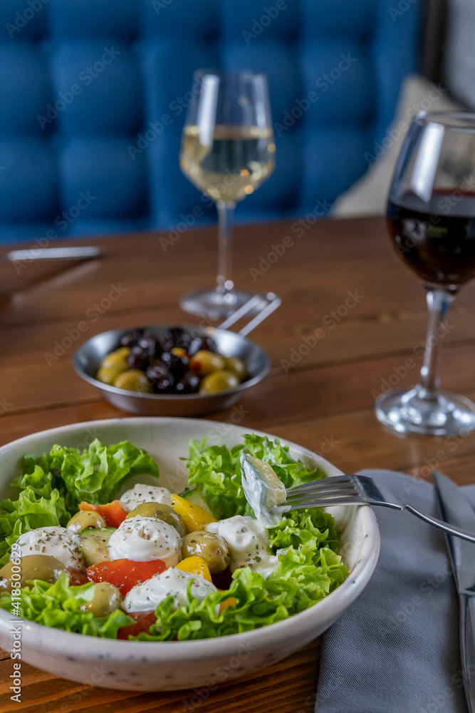 Fresh vegan green greek salad with green leaves mix, olives and vegetables. Enjoying tasty vegan food with glass of wine with friends in restaurant. Selective focus