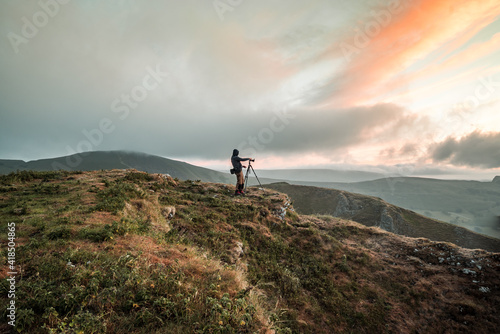 Photographer on top summit of mountain hill at dawn as sunrise illuminates clouds in sky beautiful orange. Using a tripod in rural landscape of Peak District taking photograph. 