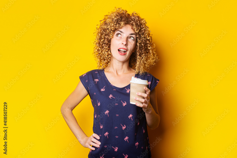 surprised young woman with curly hair holding paper cup on yellow background. surprised young woman with curly hair holding paper cup on yellow background.