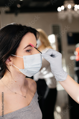 The professional eyebrow master is making an eyebrow correction for the girl in a medical mask in beauty salon. A beauty salon services during a pandemic.