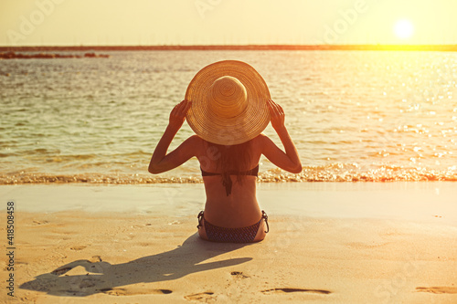Young girl at sunset on the beach sits with her back to the sea in a hat.