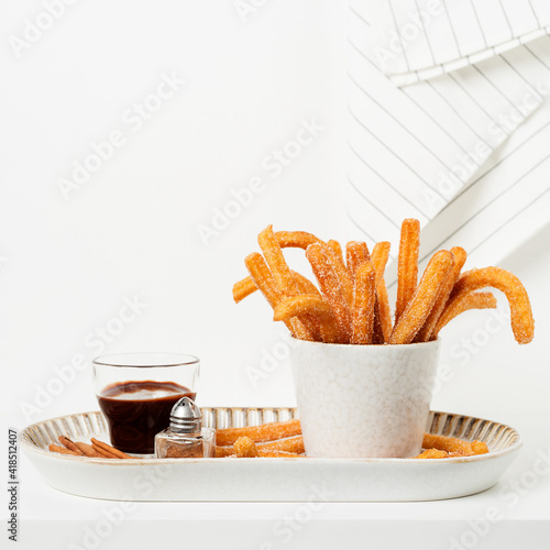 Churro sticks with sugar and chocolate sauce in kitchen shelf. White wall 