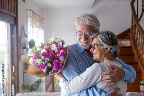 Portrait of couple of two happy and in love seniors or mature and old people holding flowers at home looking outside. Pensioners adult enjoying and celebrating holiday together.