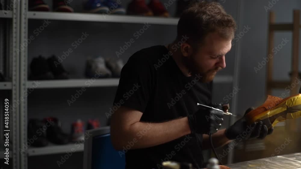 Tracking shot of professional shoemaker wearing black gloves spraying paint of light brown leather shoes. Process of repairing and restoration shoes by cobbler in shoe repair shop with dark interior. Tracking shot of professional shoemaker wearing black gloves spraying paint of light brown leather shoes. Process of repairing and restoration shoes by cobbler in shoe repair shop with dark interior.