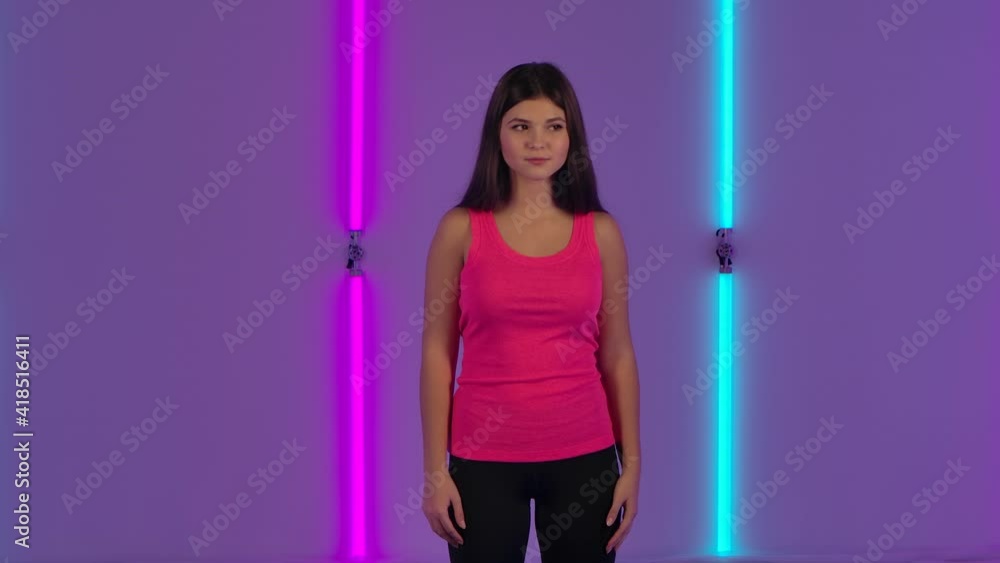 Portrait of fashionable model coquettishly looking at camera. Young brunette woman with long hair in a pink t-shirt poses against background of bright neon lights in dark studio. Slow motion. Portrait of fashionable model coquettishly looking at camera. Young brunette woman with long hair in a pink t-shirt poses against background of bright neon lights in dark studio. Slow motion.