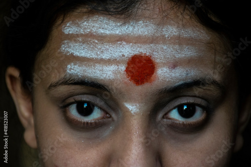 Stock photo of a beautiful 20 to 30 aged Indian hindu girl who apply horizontal white color vibhuti and round shape red color sindur tilak or vermillion tilak on her forehead looking at camera. photo